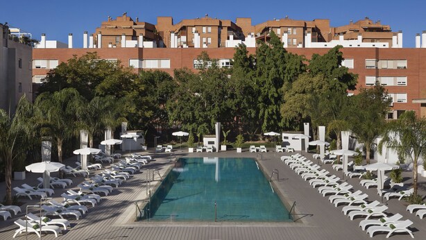 Piscina con sillones de descanso y sombrillas, rodeada de árboles y edificios.