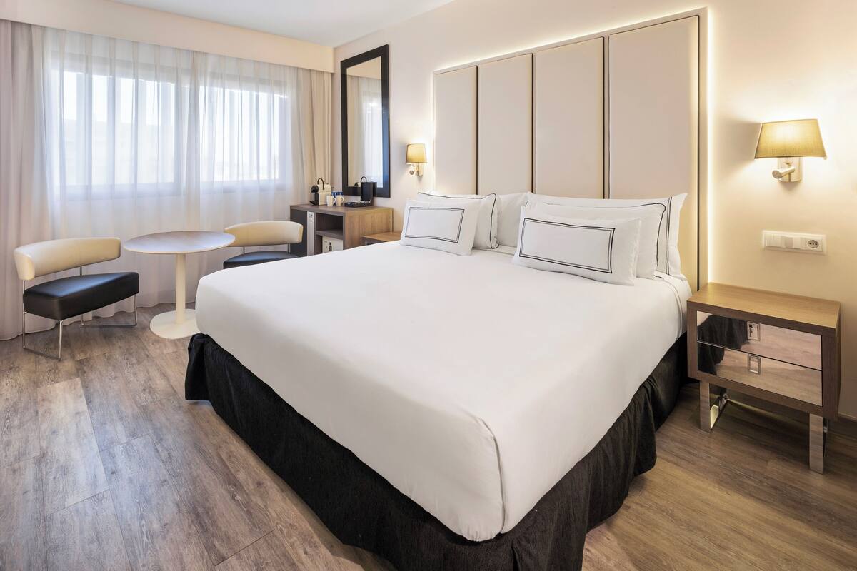 A hotel room with a large bed, a desk, a chair, and a small table.