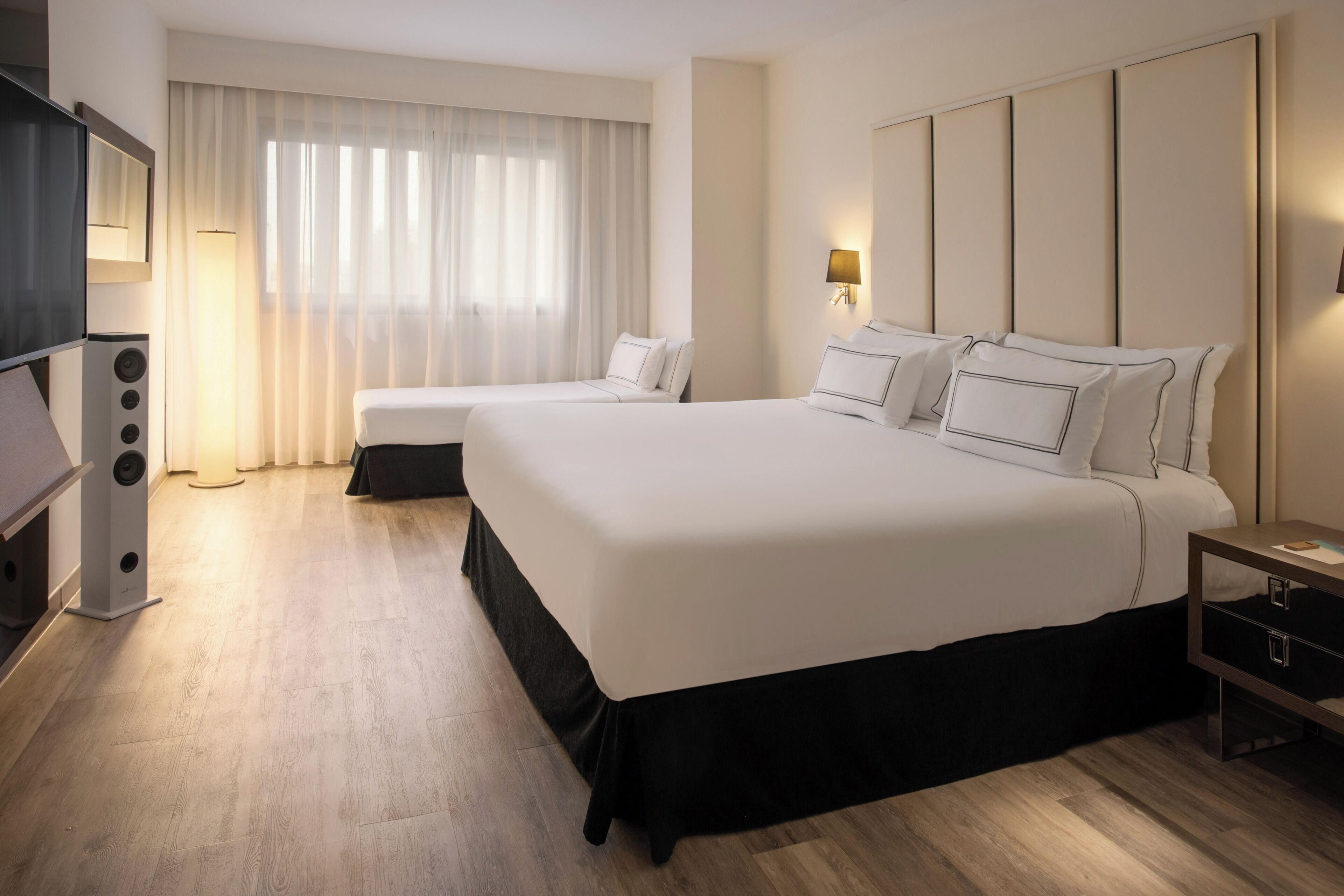 Habitación de hotel con dos camas, televisión, lámpara y un ventanal grande con cortinas.