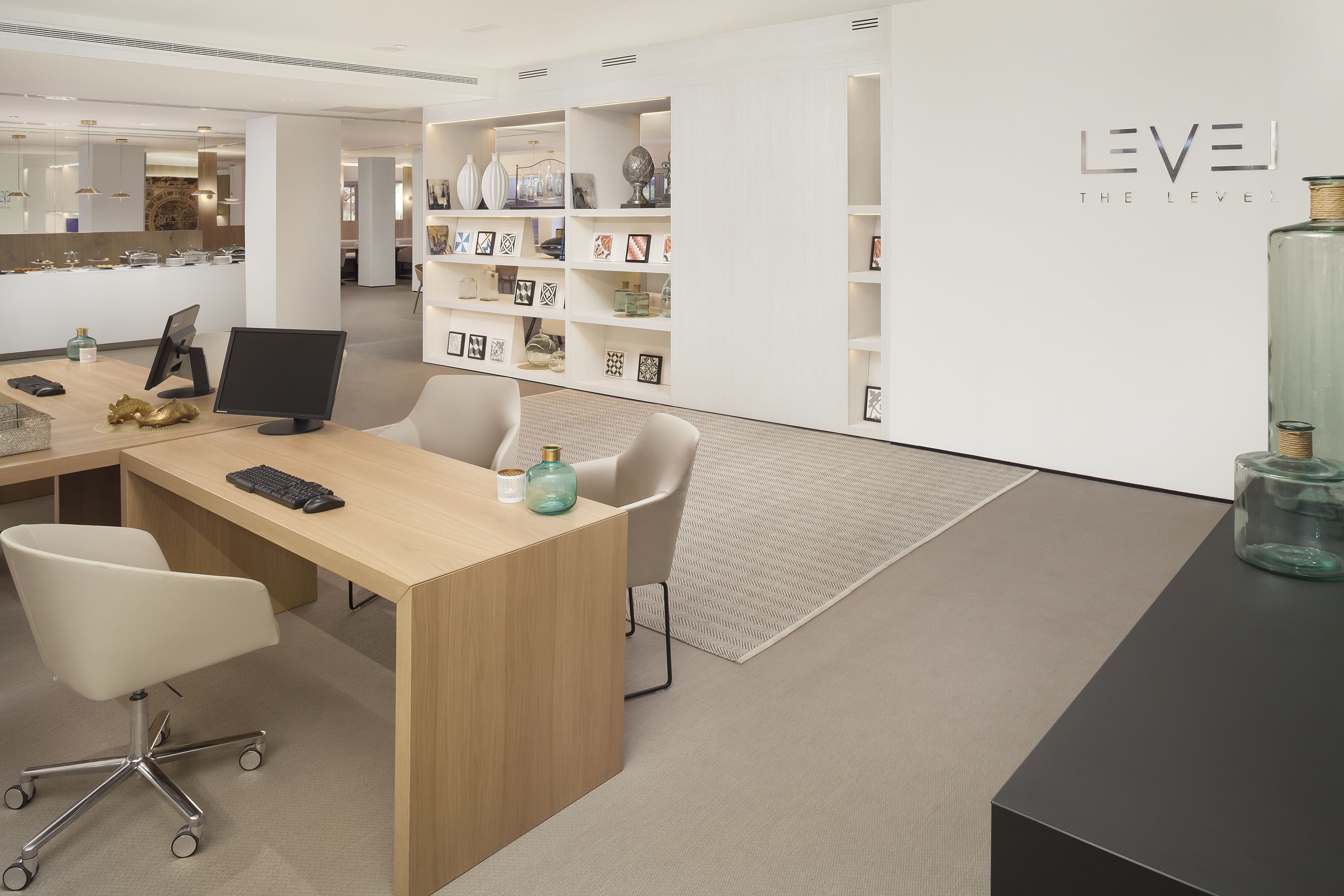 A modern office space with a wooden desk, two chairs, a computer monitor, and a bookshelf filled with decorative items.