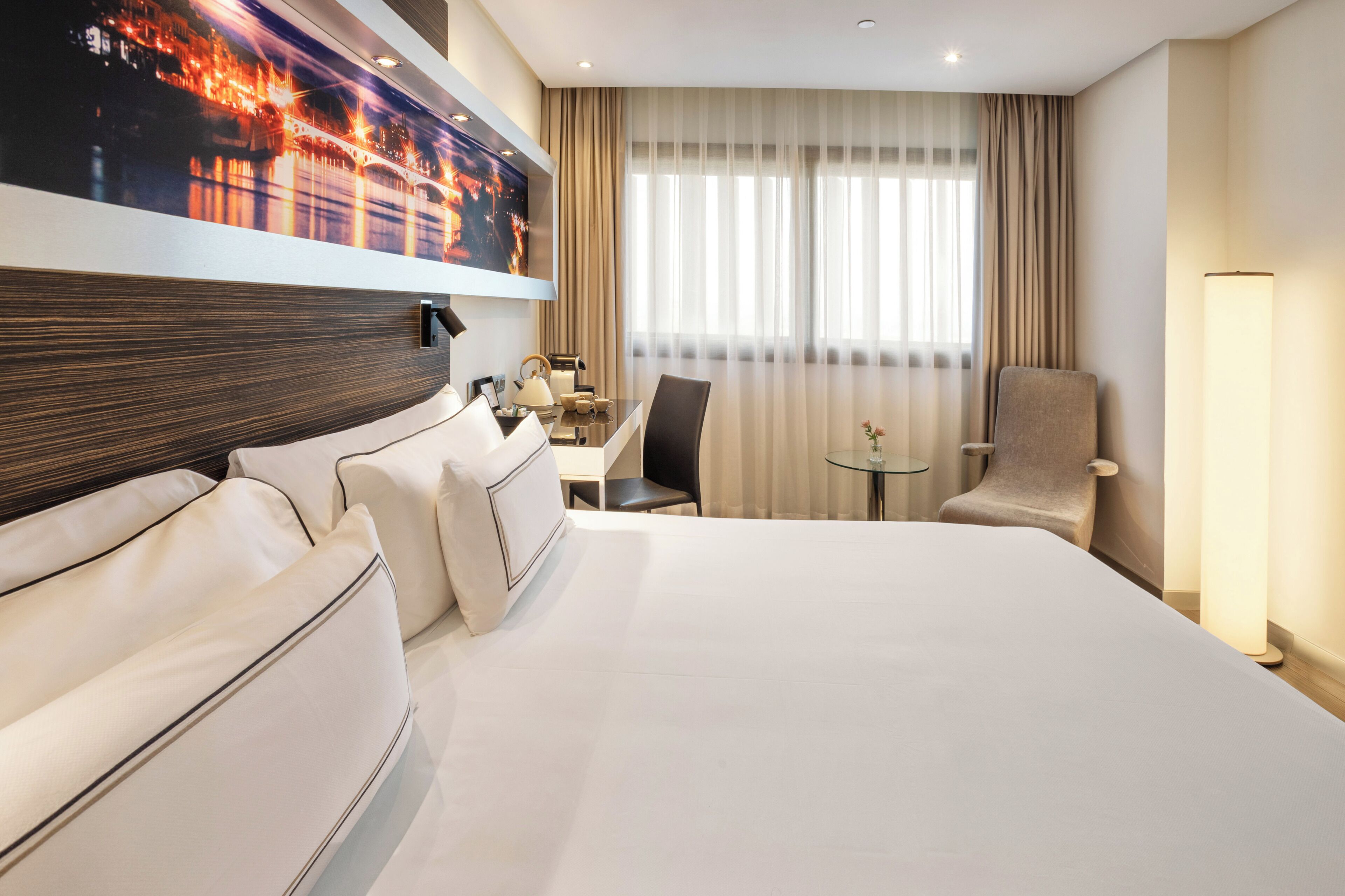 Habitación de hotel con una cama grande, un escritorio, una silla, un televisor montado en la pared y una ventana con cortinas.