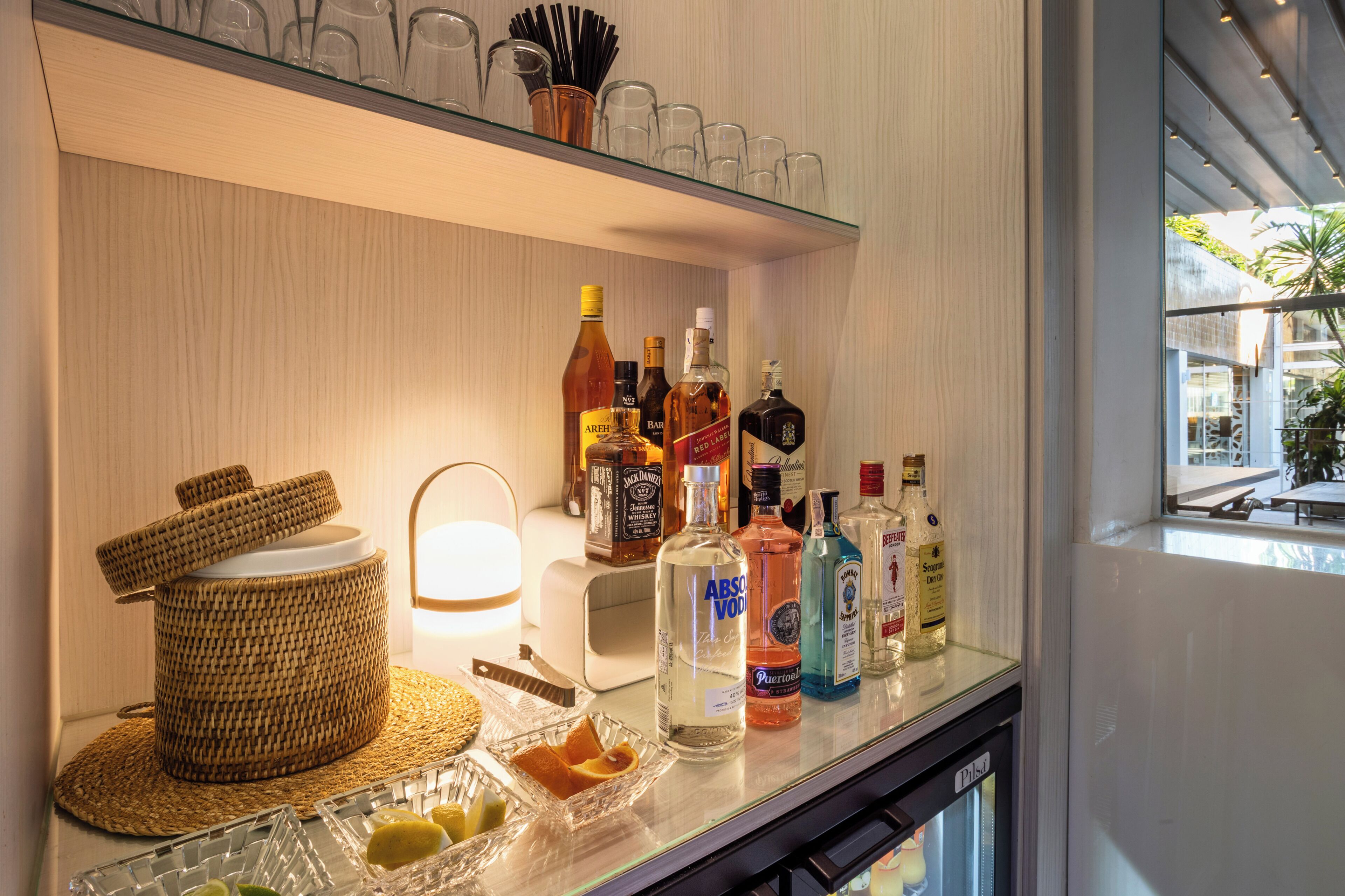 A bar area with a glass shelf holding various bottles, a woven basket, and a lit lamp.