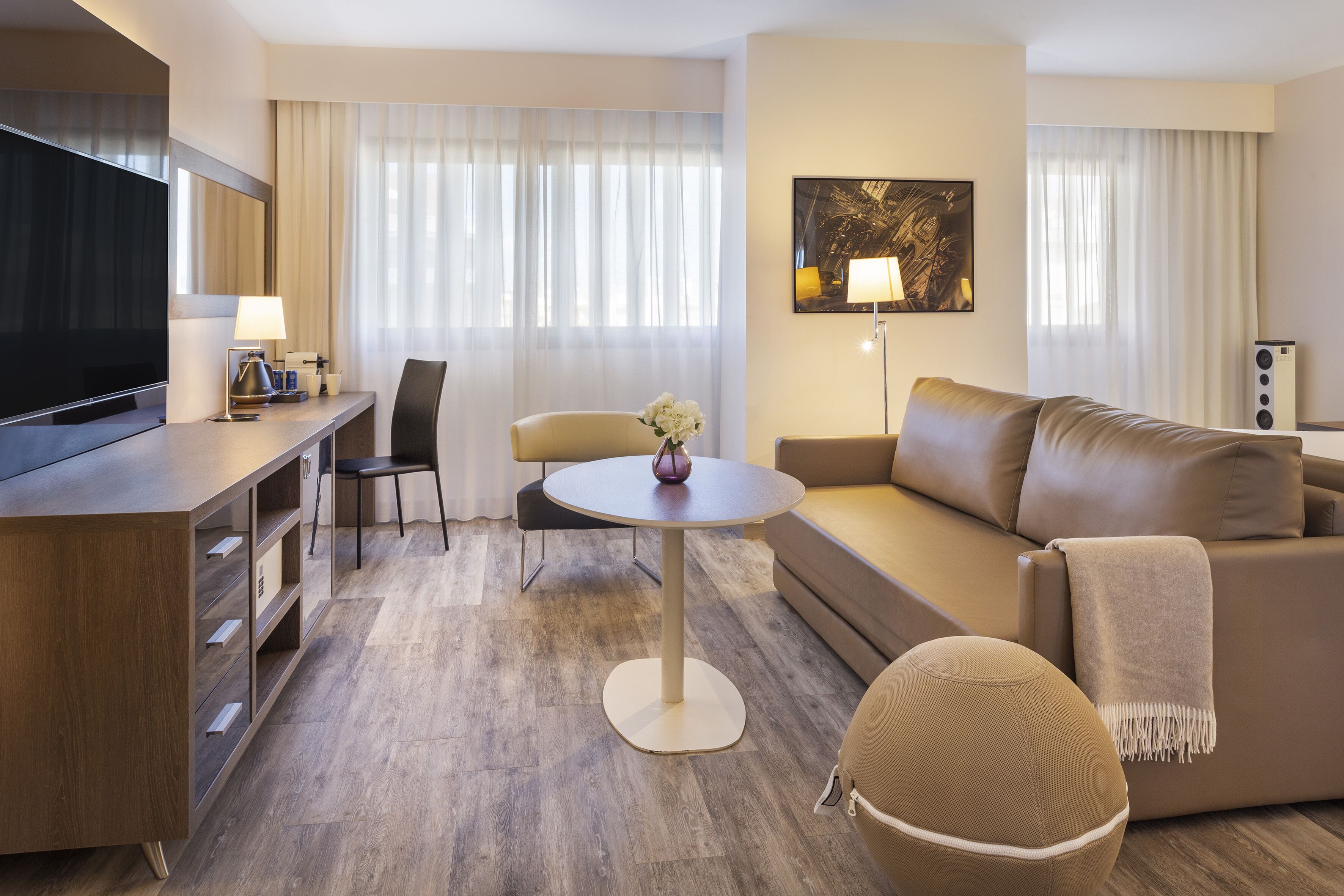 Una habitación de hotel moderna con una televisión de pantalla plana, una mesa redonda para comer, un sofá, una silla y un pequeño taburete.