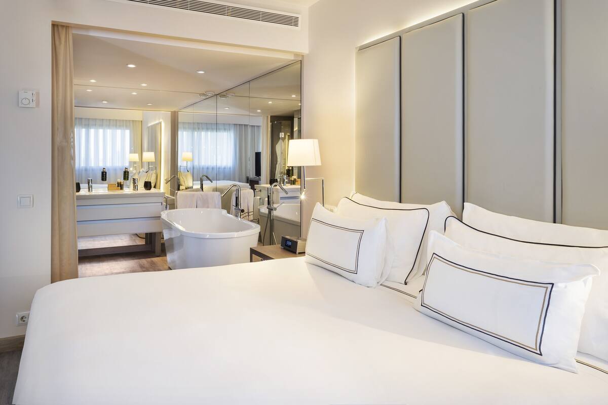 A modern hotel room with a large bed, a separate bathroom with a bathtub, and a mirror reflecting the room.
