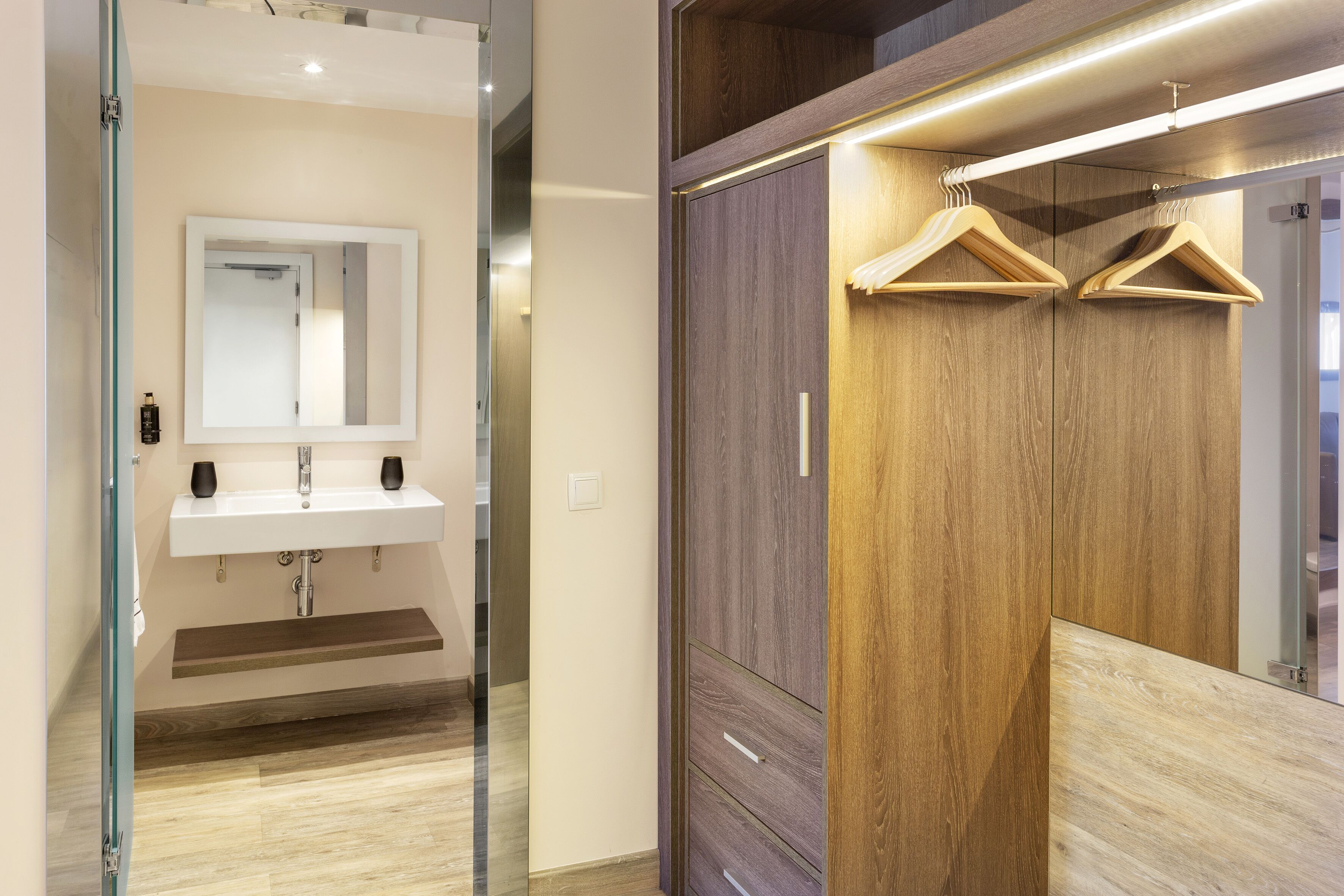 Un baño moderno con cabina de ducha de vidrio, un lavamanos blanco con dos dispensadores de jabón negros y un mueble de tocador de madera con un espejo y dos colgadores de madera.