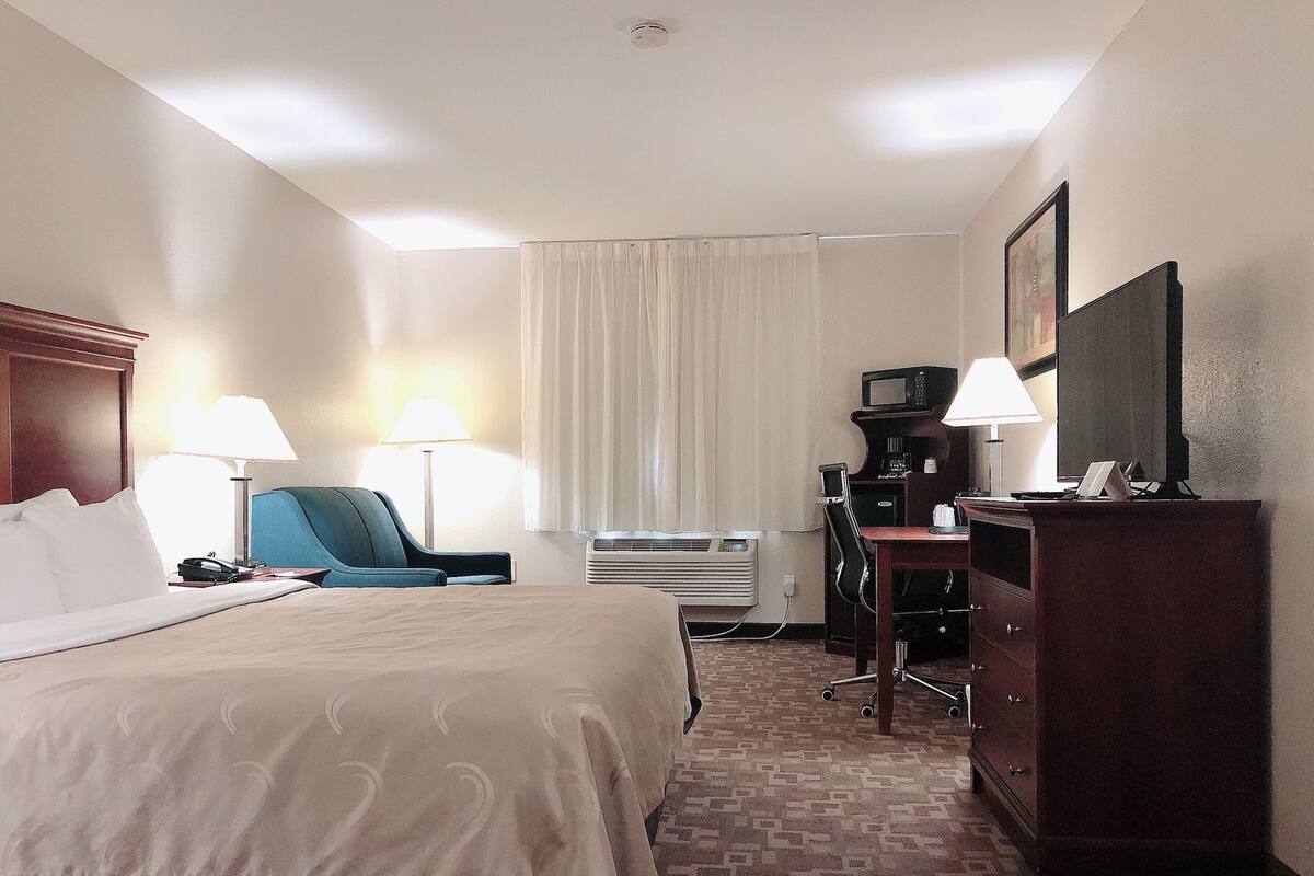 A hotel room with a bed, two chairs, a desk with a computer, a TV, and an air conditioning unit.