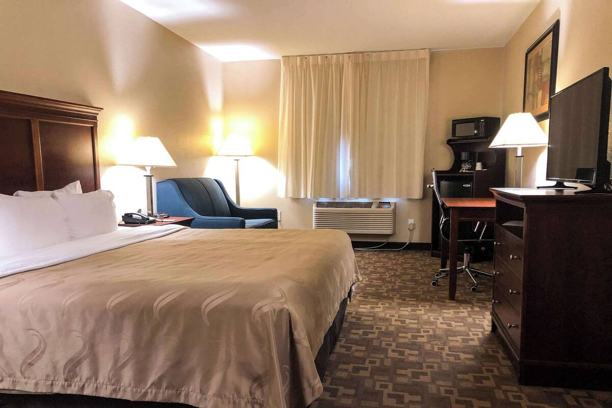 A hotel room with a large bed, two bedside lamps, a desk with a computer, a chair, and a dresser.