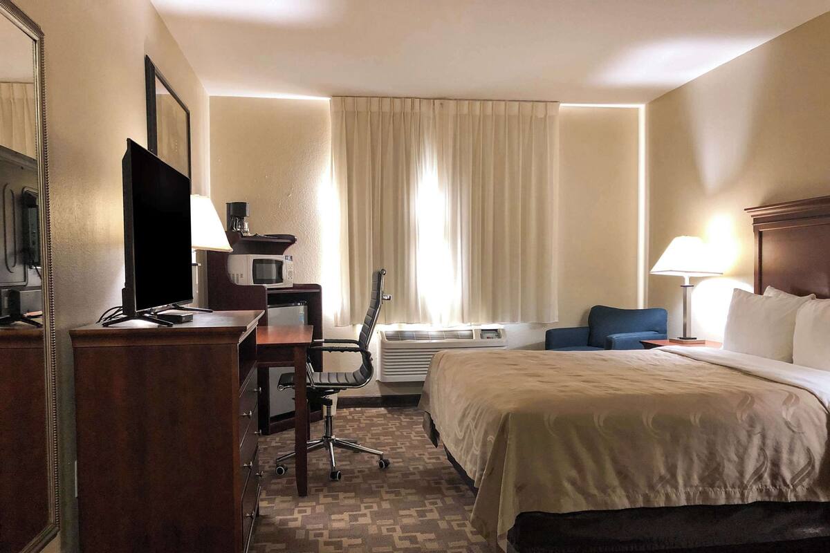 A hotel room with a bed, desk, chair, TV, lamp, and air conditioning unit.