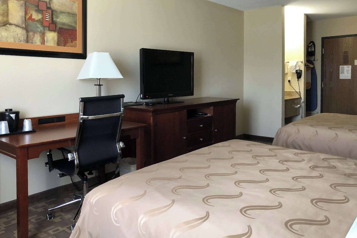 A hotel room with a desk, a chair, a television, a bed with patterned bedding, and a door leading to another room.