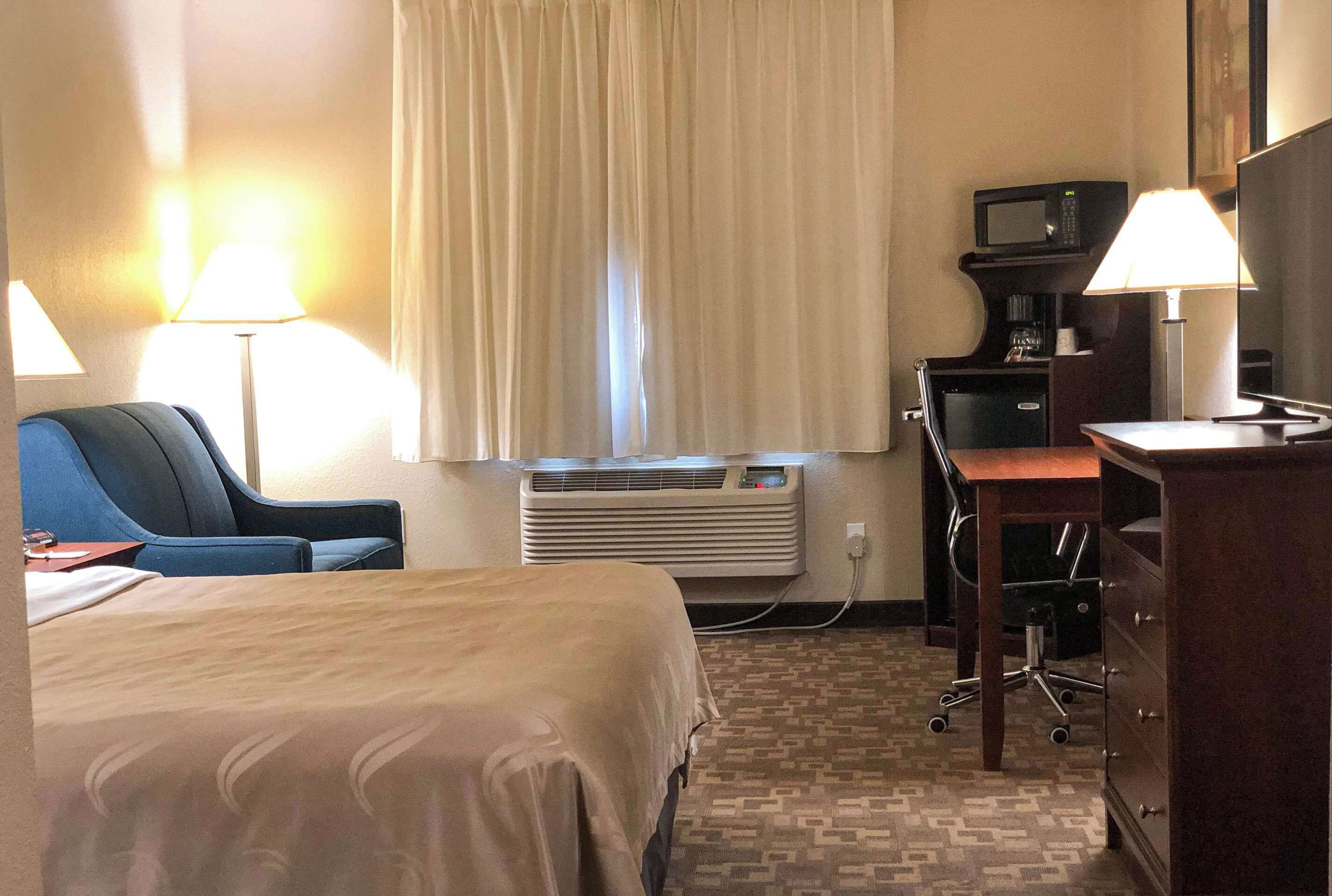 A hotel room with a bed, a desk, a chair, a desk lamp, a television, a microwave, and an air conditioning unit.