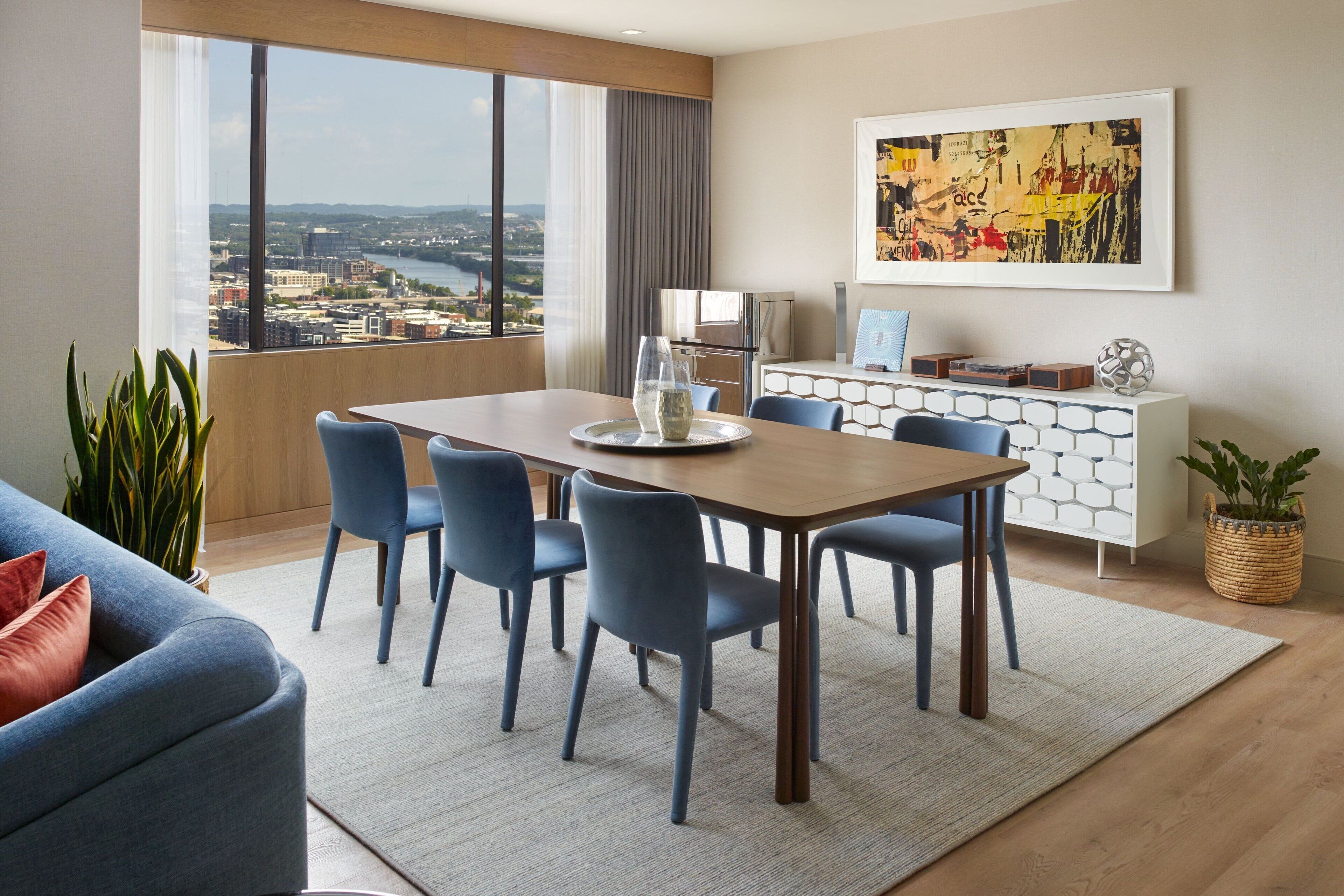 Habitación de hotel moderna con zona de comedor, ventanas amplias con vista a la ciudad, sofá y un elemento decorativo en la pared.