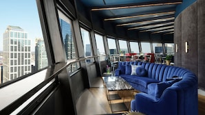 Ein modernes Interieur mit einem blauen, gebogenen Sofa, einem Glastisch und Blick auf die Stadt durch große Fenster.