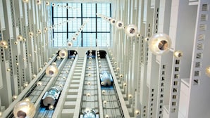 Ein modernes Interieur mit einer zentralen Rolltreppe, flankiert von zylindrischen Säulen, einem großen Fenster am Ende und einer Reihe kugelförmiger Leuchten.