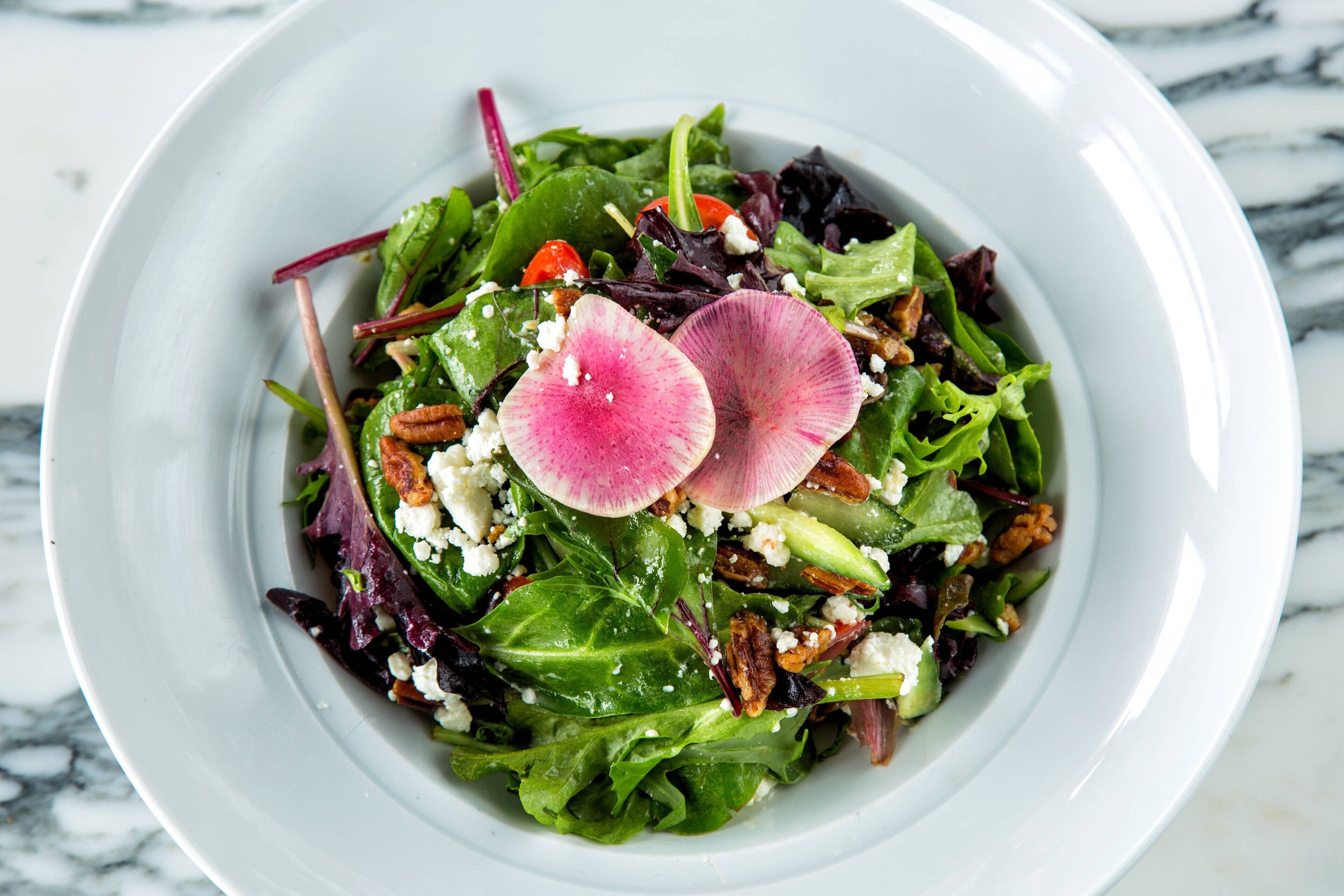 A white plate with a salad containing mixed greens, nuts, and crumbled cheese, garnished with radish slices.