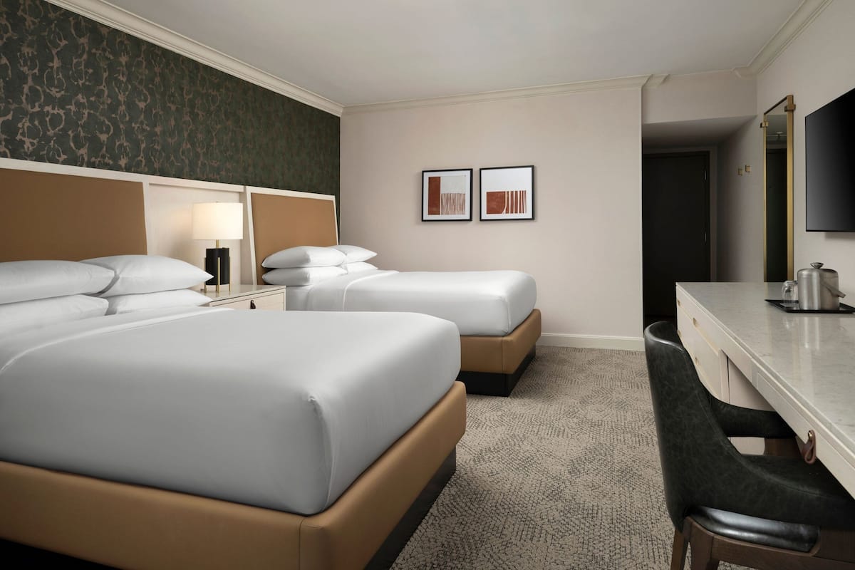 Une chambre d’hôtel avec deux lits, un bureau, une chaise, une lampe et deux tableaux encadrés au mur.