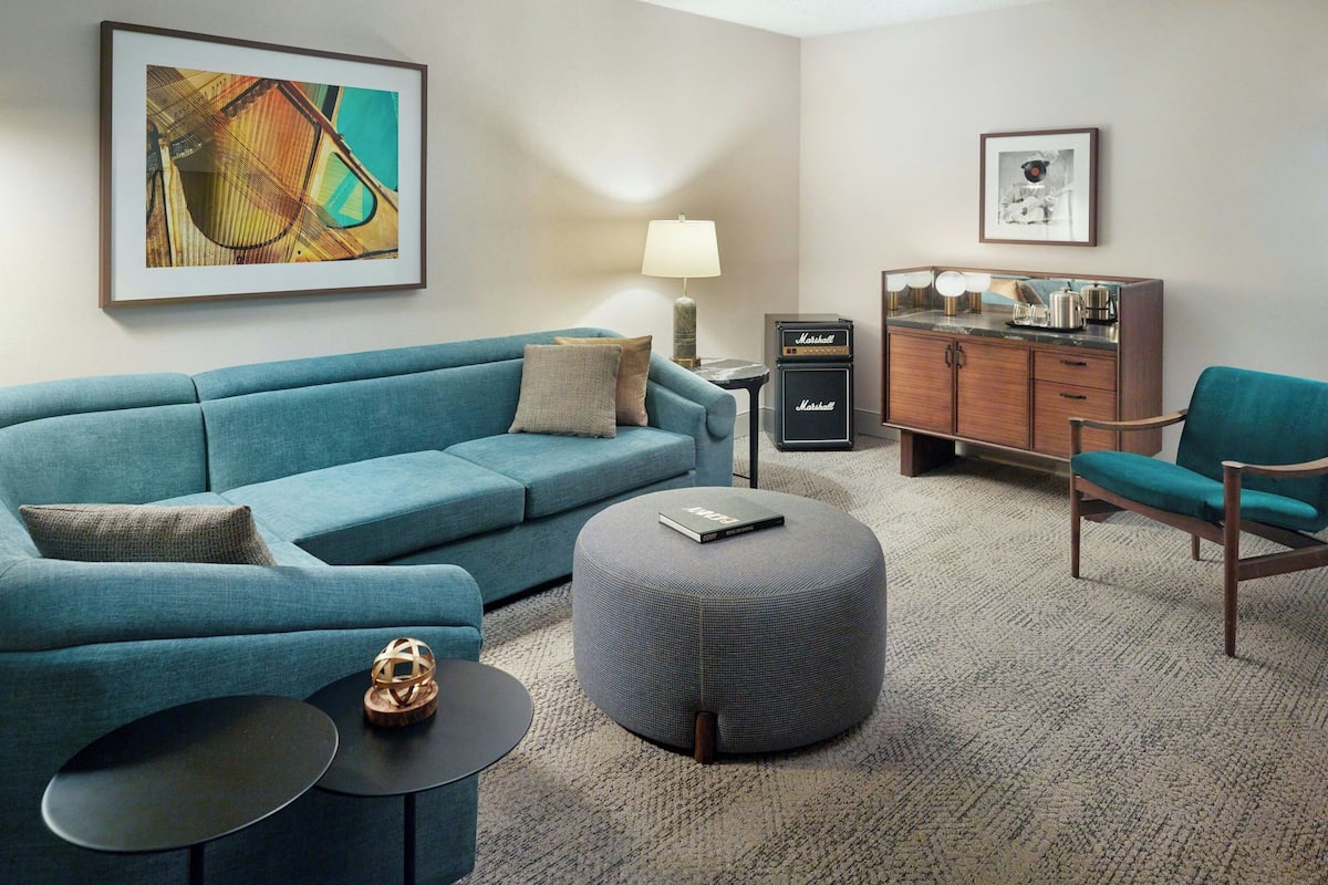 Un salon moderne avec un canapé bleu, un pouf rond, une petite table d’appoint, une lampe, un meuble avec un miroir et des œuvres d’art encadrées accrochées aux murs.