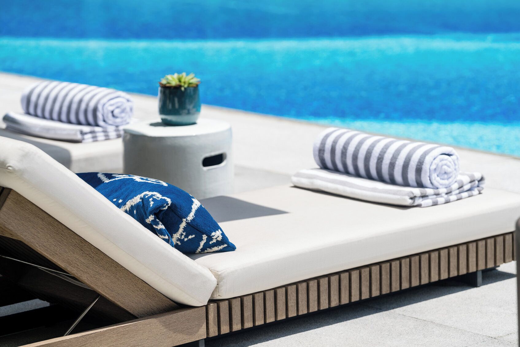 A sun lounger with a rolled towel and a blue pillow, a small potted plant, and a striped towel on a poolside area.