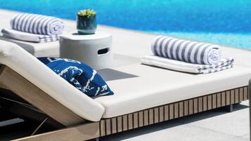 A sun lounger with a rolled towel and a blue pillow, a small potted plant, and a striped towel on a poolside area.