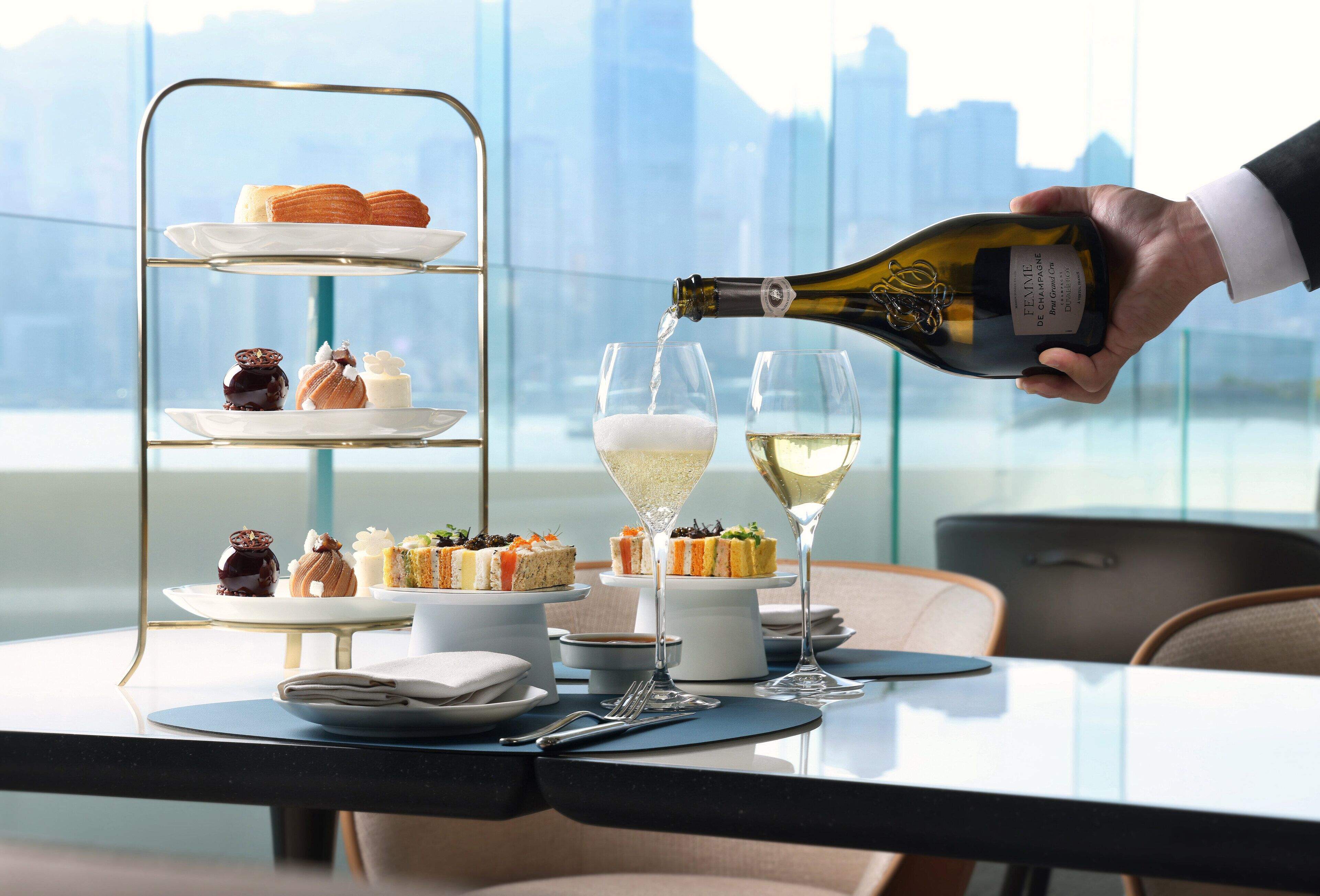 A three-tiered dessert stand with various pastries and cakes, a bottle of champagne being poured into two glasses, and a cityscape view in the background.