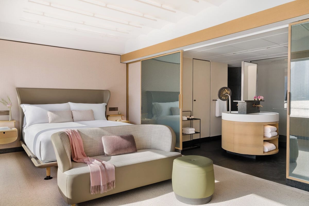 Une chambre d’hôtel moderne dotée d’un grand lit, d’un canapé, d’un pouf rond, d’une salle de bain avec un meuble-lavabo et un miroir, et d’un placard à portes coulissantes.