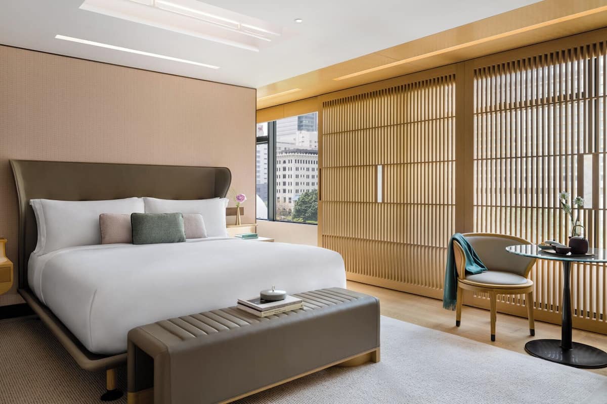Une chambre d’hôtel moderne avec un grand lit, une table de chevet, une chaise, une petite table avec une théière, et une vue sur la ville grâce à de grandes fenêtres.