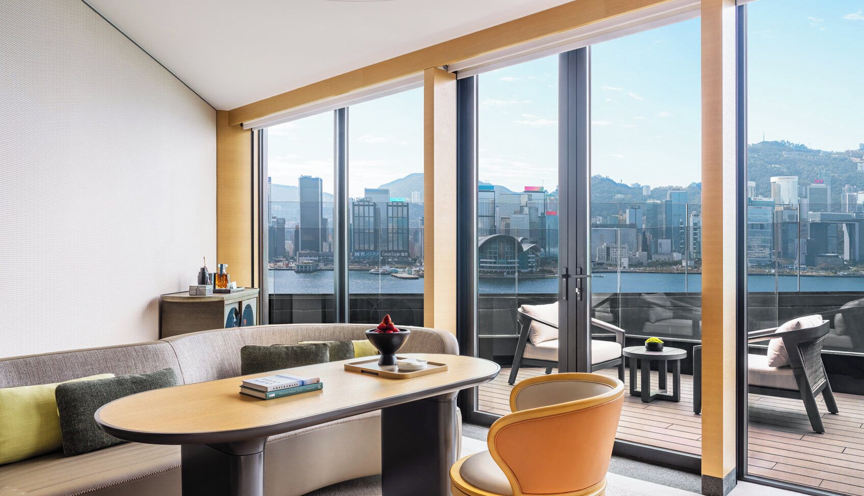 A modern hotel room with a large balcony, a sofa, a dining table, and a view of a cityscape.