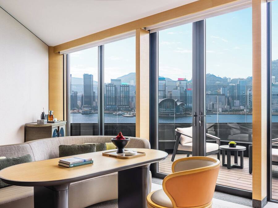 A modern hotel room with a large balcony, a sofa, a dining table, and a view of a cityscape.