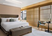 A modern hotel room with a large bed, a bedside table, a chair, a small table with a teapot, and a view of the city through large windows.