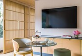 A modern room with a glass table, a sofa, a TV mounted on the wall, and a window with a view of greenery.