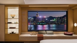 A modern hotel room with a large window offering a nighttime cityscape view, a cozy seating area, and a built-in bookshelf. - Regent Hong Kong (Kowloon)