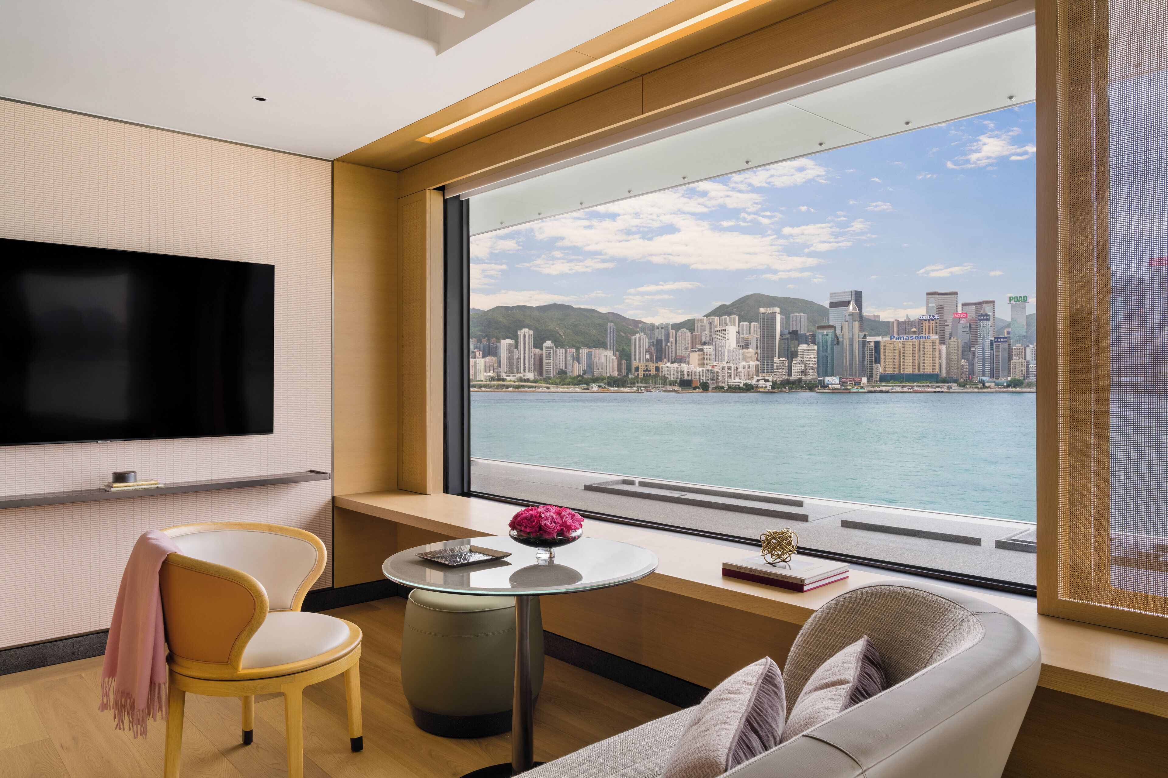 Habitación de hotel moderna con un amplio ventanal que ofrece vistas a la silueta de la ciudad, un televisor de pantalla plana montado en la pared, un sofá cómodo y una pequeña mesa redonda con un florero.