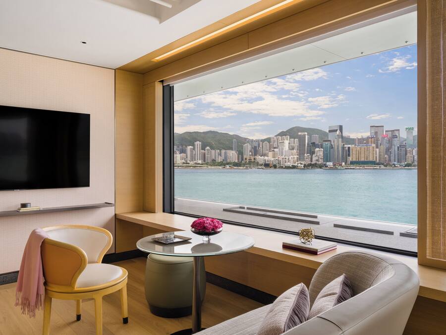 Habitación de hotel moderna con un amplio ventanal que ofrece vistas a la silueta de la ciudad, un televisor de pantalla plana montado en la pared, un sofá cómodo y una pequeña mesa redonda con un florero.