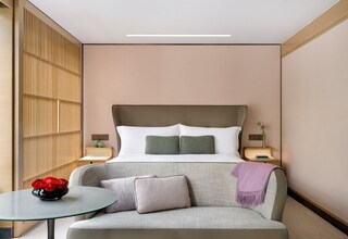 A modern hotel room with a large bed, a grey sofa, a small round table with a vase of red flowers, and a decorative wall panel.