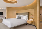 A modern hotel room with a curved headboard, a flat-screen TV mounted on the wall, a bedside table with a vase of flowers, and a built-in wardrobe.