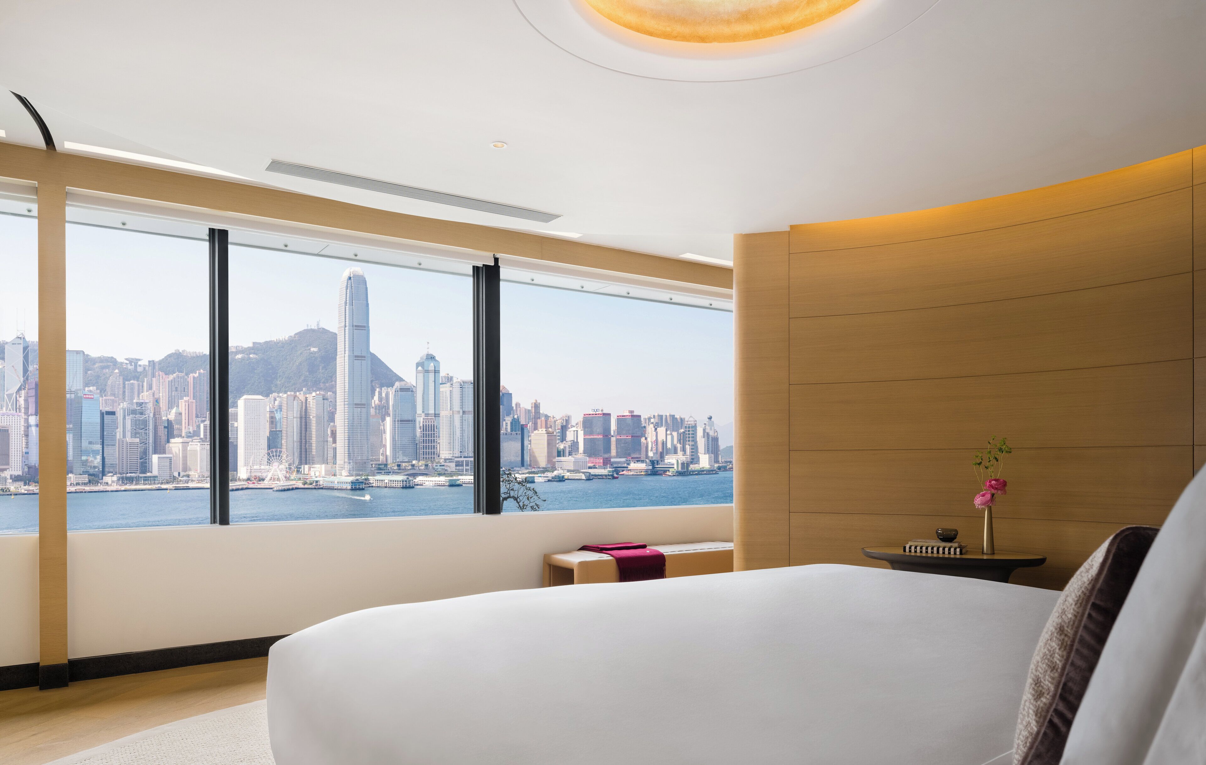 Habitación de hotel moderna con un ventanal que ofrece vistas a un horizonte urbano, una cama, una mesita de noche con un jarrón de flores y un panel de pared de madera.