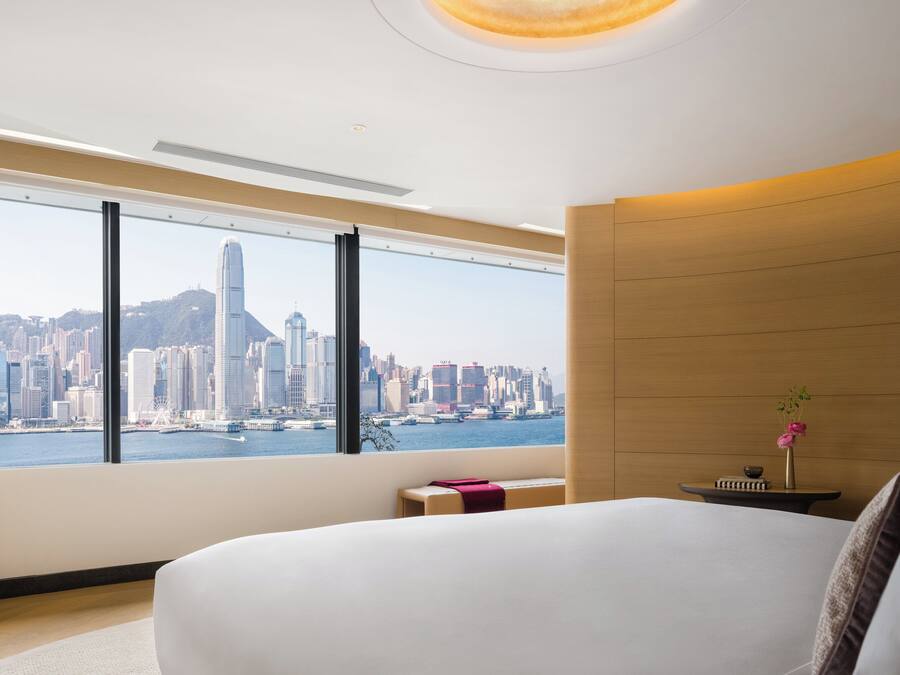 Habitación de hotel moderna con un ventanal que ofrece vistas a un horizonte urbano, una cama, una mesita de noche con un jarrón de flores y un panel de pared de madera.