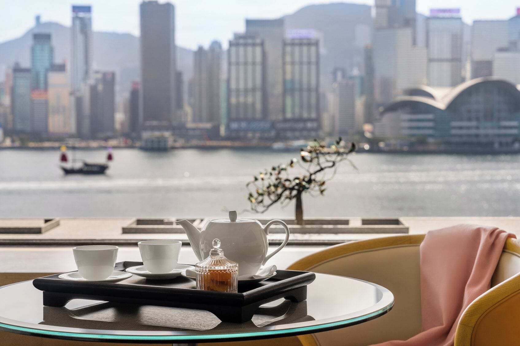 A table with a tray holding a teapot, cups, and a small container, set against a view of a city skyline and a river.