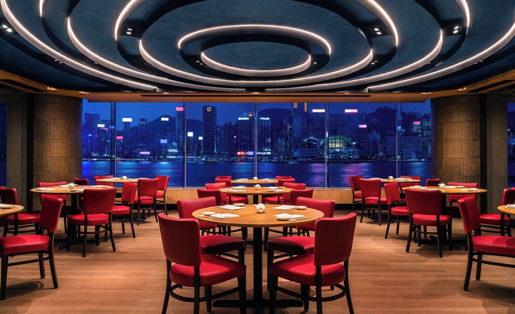 A restaurant with round tables and red chairs, large windows with a city view, and a modern ceiling design.