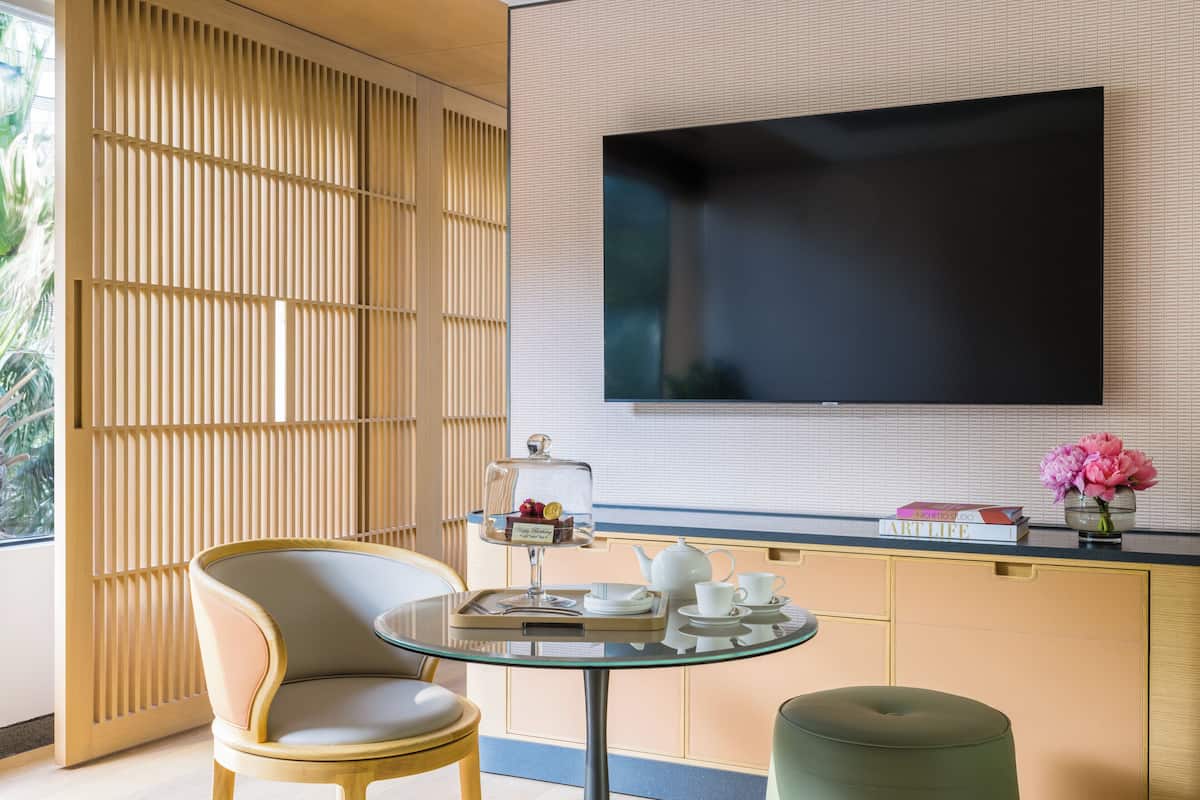 Une pièce moderne avec une table en verre, un canapé, un téléviseur fixé au mur et une fenêtre donnant sur un paysage verdoyant.