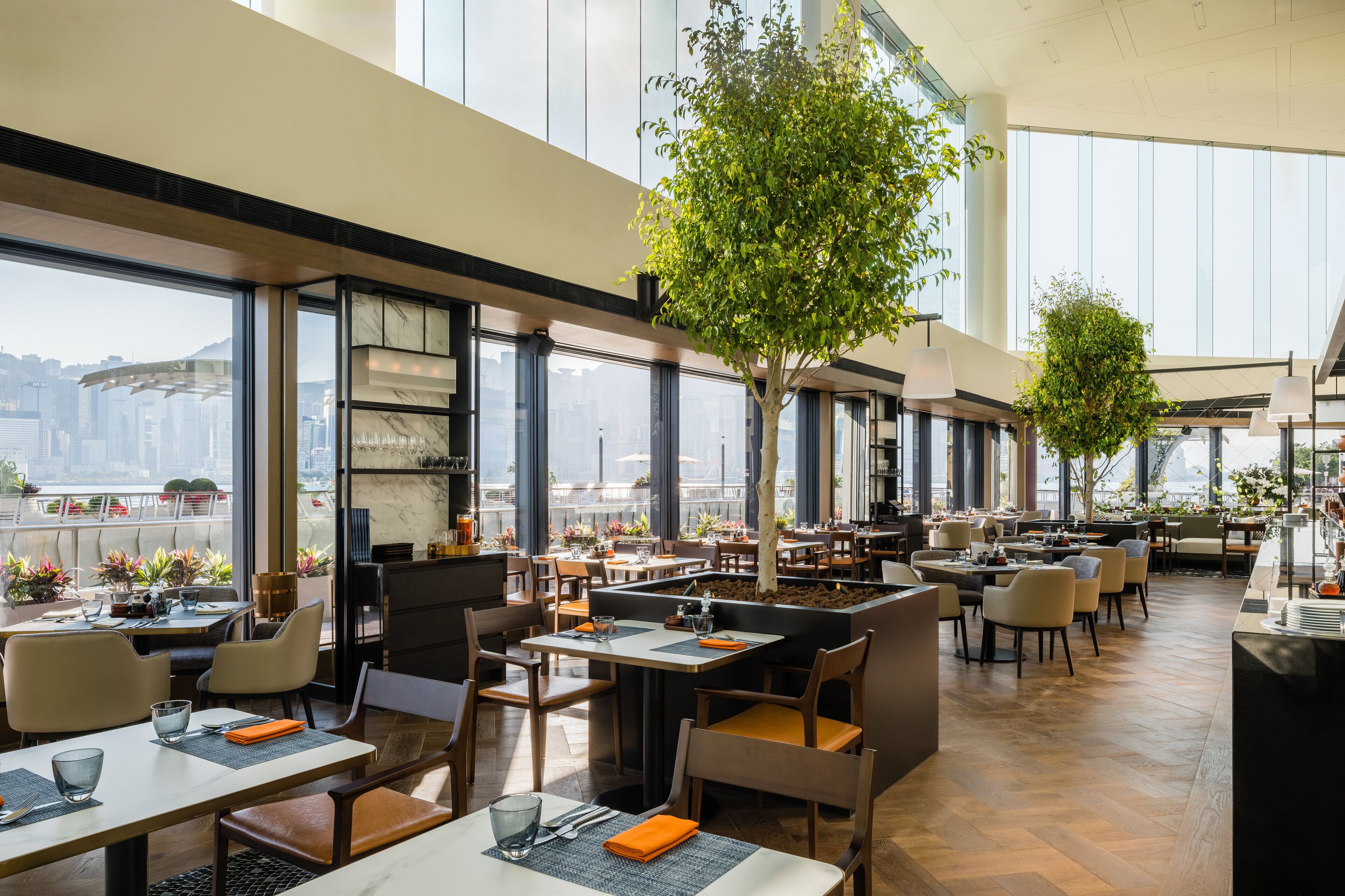 A spacious restaurant with high ceilings, large windows, and indoor trees. Tables are neatly arranged with chairs.