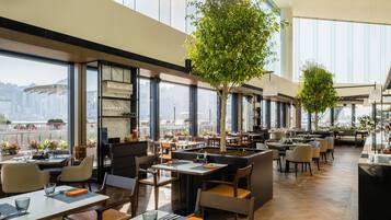 A spacious restaurant with high ceilings, large windows, and indoor trees. Tables are neatly arranged with chairs.