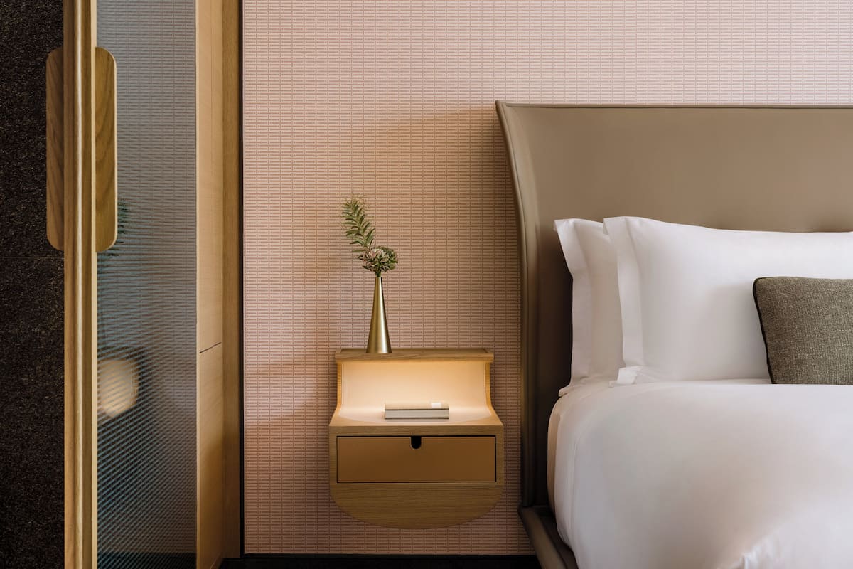 Un lit bien fait, avec du linge de lit blanc, une petite table de chevet sur laquelle se trouvent un livre et un vase de fleurs, et un mur orné d’un motif discret.