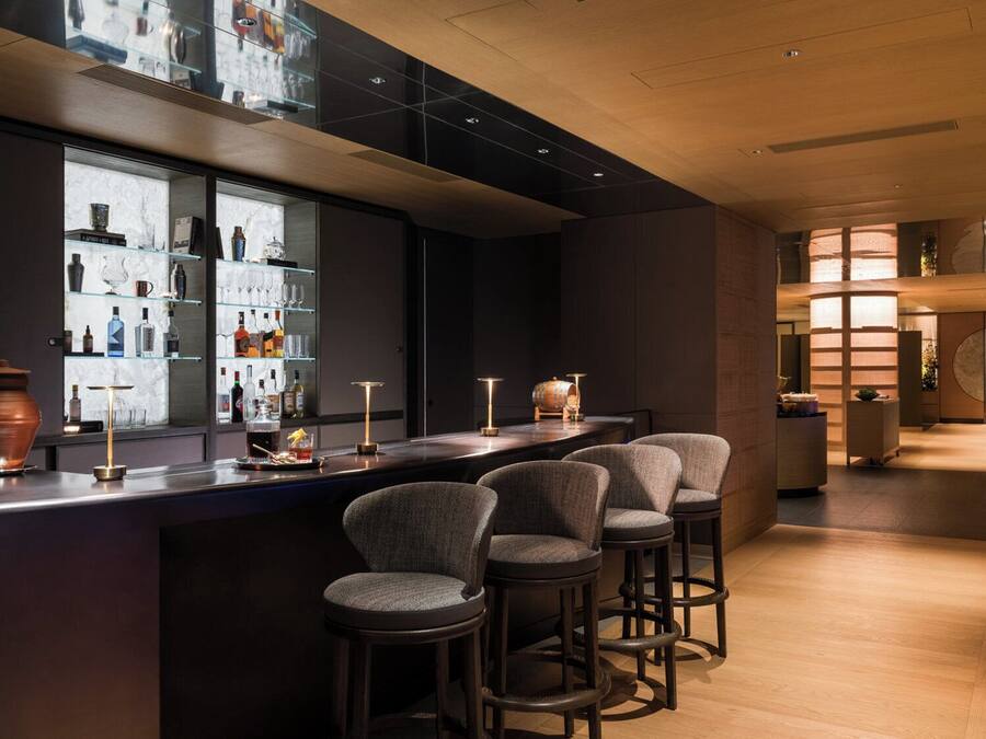 Una zona de bar con mostrador de madera oscura, varios taburetes y una vitrina con botellas y vasos.