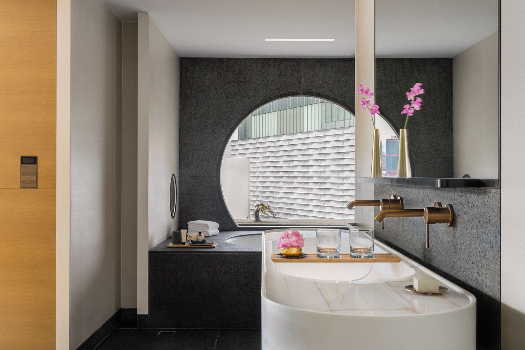 A modern bathroom with a large bathtub, a round mirror, and a view of a building through a circular window.
