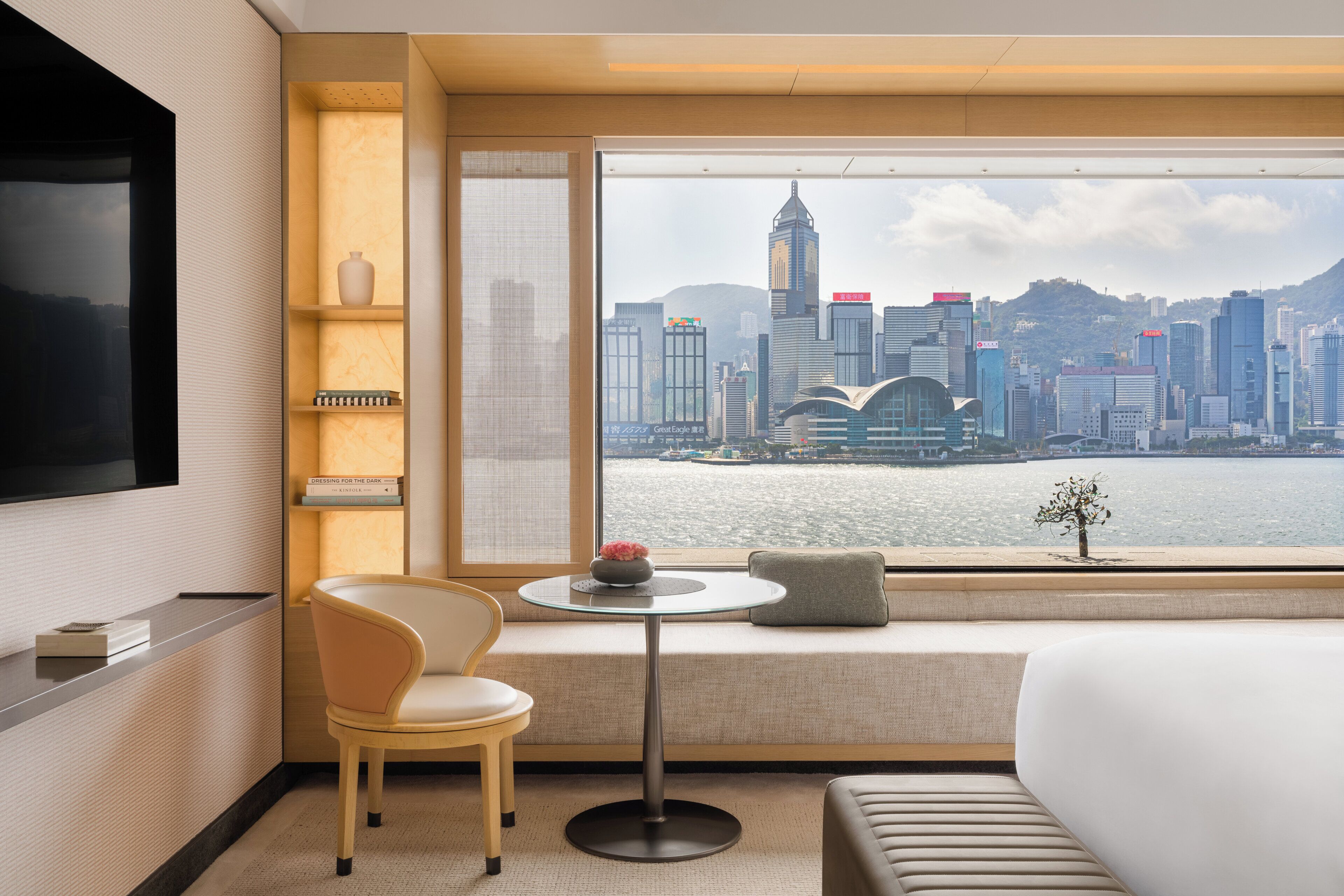 A hotel room with a large window, a bed, a TV, a chair, a small table, and a bookshelf.