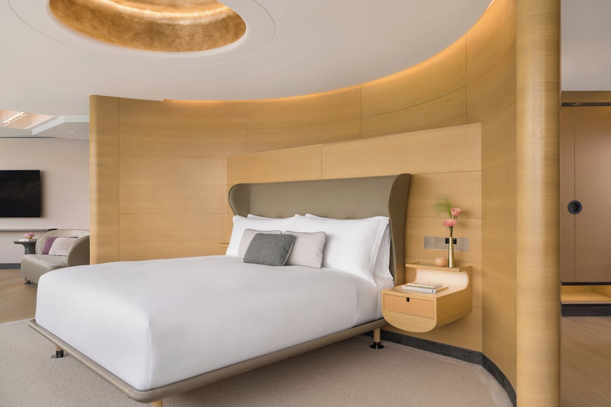 Une chambre d’hôtel moderne dotée d’une tête de lit incurvée, d’un téléviseur à écran plat fixé au mur, d’une table de chevet ornée d’un vase de fleurs et d’une petite table de nuit avec une lampe.