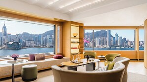 A modern hotel room with a large window offering a city skyline view, a comfortable seating area, and a small table with a vase of flowers. - Regent Hong Kong (Kowloon)