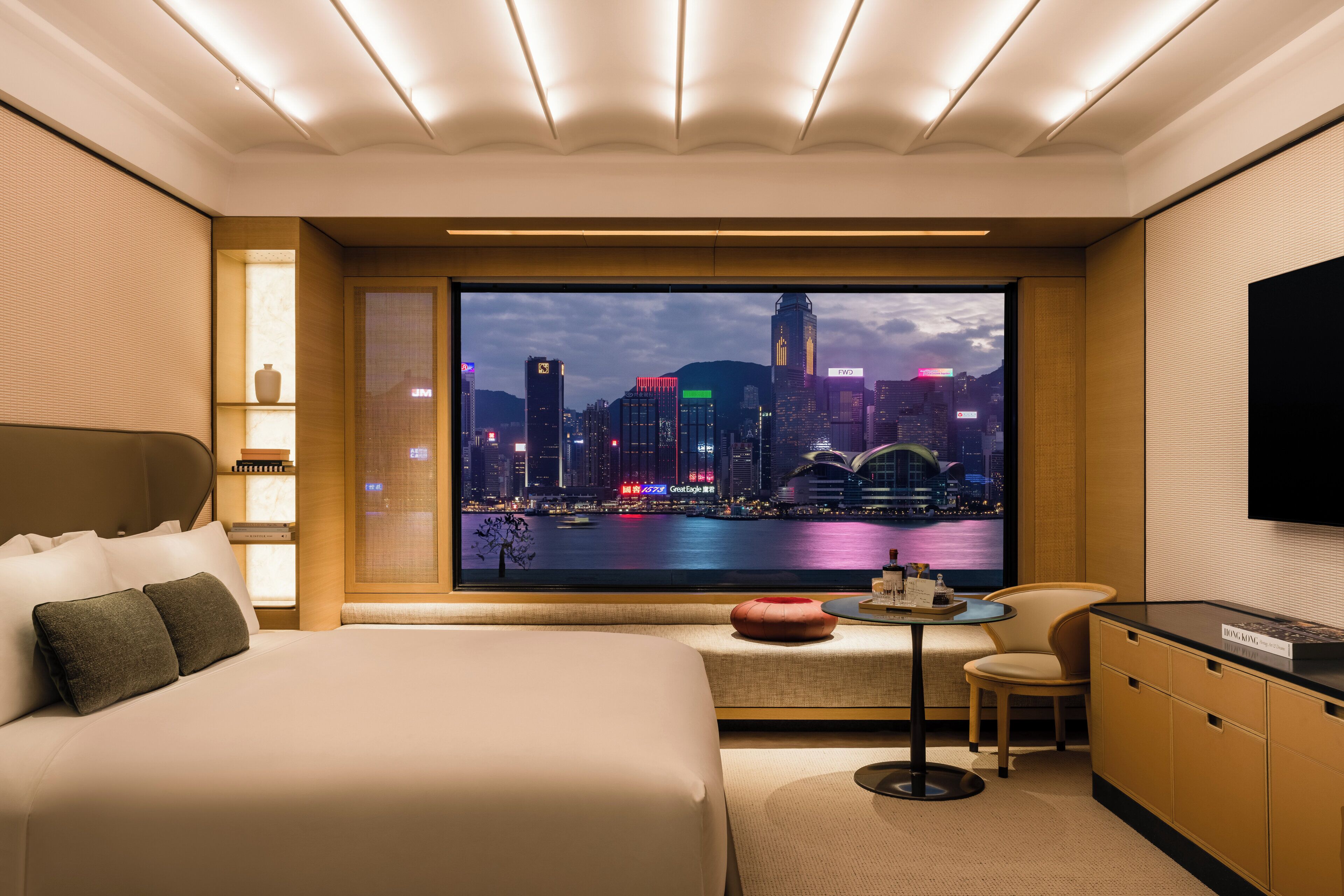Habitación de hotel con un ventanal grande que ofrece vista a la ciudad, cama con almohadas, una mesita con una silla y un televisor en la pared.