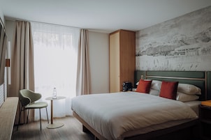 Una habitación de hotel con una cama, una silla, una mesita con una botella, un cuadro en la pared y una ventana con cortinas.