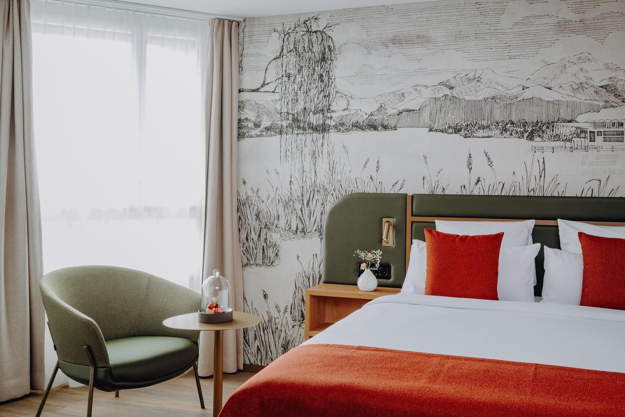 Habitación de hotel con cama, un sillón verde, una mesita con un florero y un mural de paisaje en la pared.